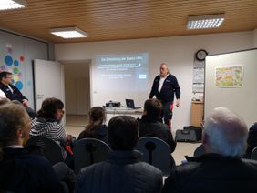 Ausbilder Peter Stock hält einen Vortrag vor einer Präsentation an der Wand. Die Zuhörenden sitzen mit dem Rücken zur Kamera.