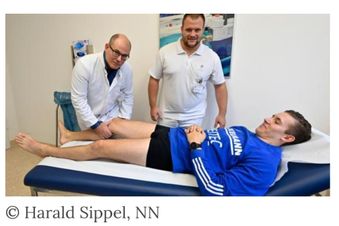 Zwei Mediziner untersuchen das Bein eines Patienten im Trikot, der auf einer Liege liegt.