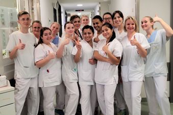 Pflegekräfte in weißer Kleidung stehen lächelnd im Flur eines Krankenhauses und zeigen Daumen hoch.