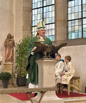 Ein Bischof steht in einer Kirche am Pult, während zwei Messdiener neben ihm sitzen.