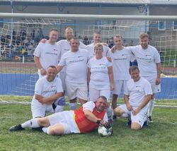 Fußballmannschaft mit neun Spielenden in weißen Trikots und einem Torwart in rot-gelbem Trikot vor einem Tor auf dem Spielfeld.