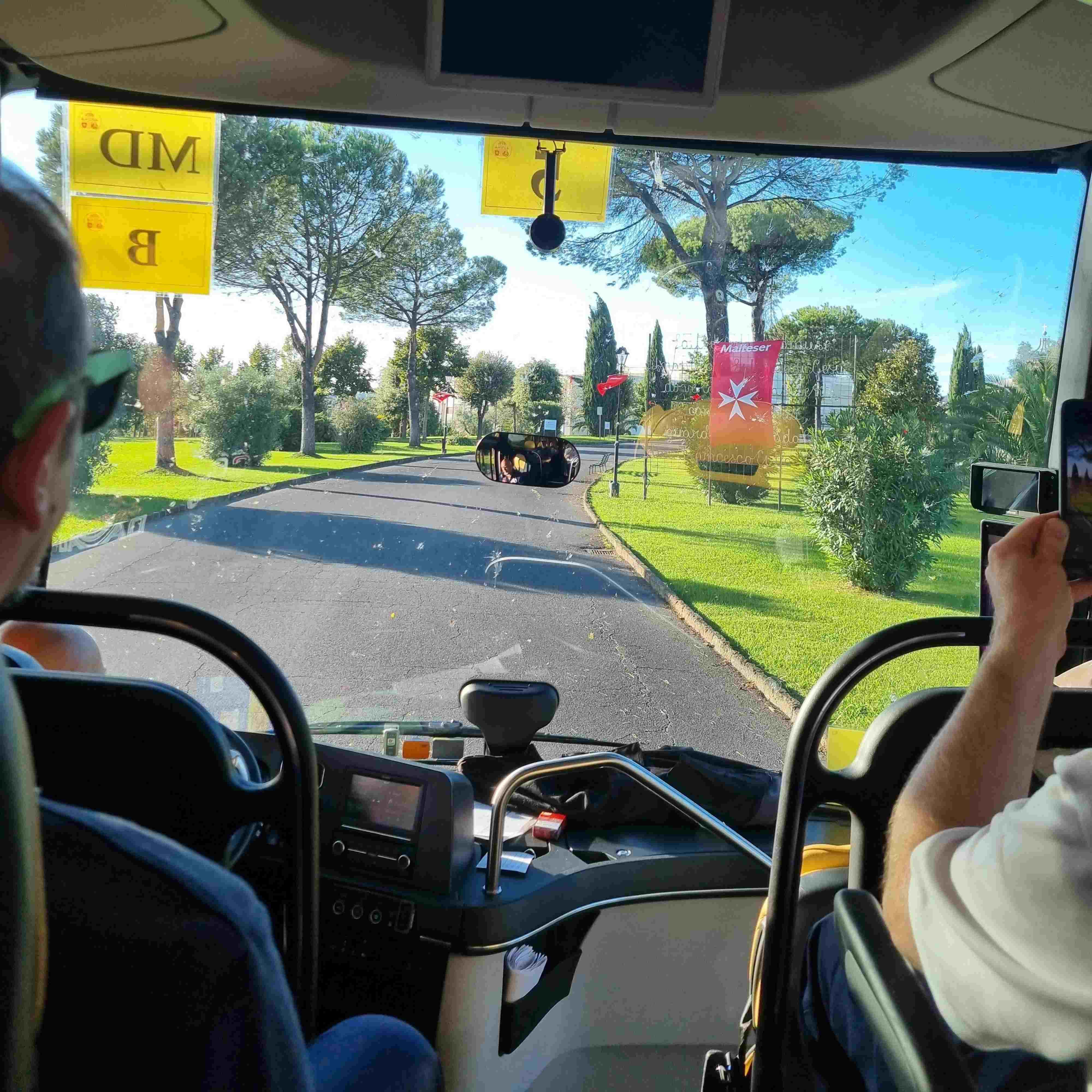 Blick aus dem Busfahrerbereich auf eine Straße mit Bäumen und roten Fahnen, zwei Personen sitzen vorne, eine hält ein Smartphone.
