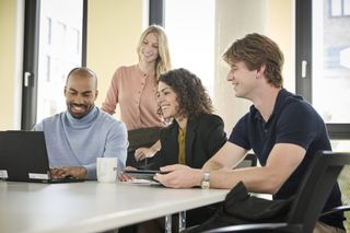 Malteser-Trainees bei einem Teammeeting am Tisch mit Laptop, engagiert und lächelnd in heller Büroumgebung
