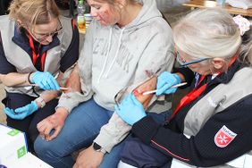 Eine Person sitzt auf einem Stuhl und bekommt von zwei Einsatzkräften Wunden geschminkt. Die Einsatzkräfte tragen blaue Einmalhandschuhe und schminken Verletzungen an beiden Armen. An Kleidung und Gesicht der sitzenden Person sind Wunddarstellungen zu sehen.
