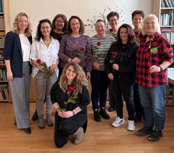 Eine Gruppe von Personen steht in einem Raum vor einer Bücherwand zusammen und hält jeweils eine einzelne rote Rose in der Hand. Eine Person kniet in der ersten Reihe, die übrigen stehen dahinter in zwei Reihen. Der Raum hat einen Holzfußboden, Regale mit Büchern und Ordnern sowie einen schlichten, hellen Hintergrund.