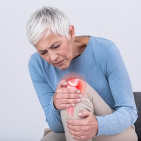 Seniorin hält sich mit schmerzverzerrtem Gesicht das Knie, rotes Symbol markiert Schmerzstelle.
