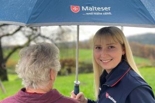 Malteser Mitarbeiterin hält einen Regenschirm über eine Seniorin