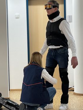 Gudrun Preßler hilft dem Freiwilligen, den Alterssimulationszug anzuziehen.