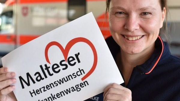 Malteser Mitarbeiterin lächelt in die Kamera und hält ein Herzenswunsch-Krankenwagen-Schild hoch. 