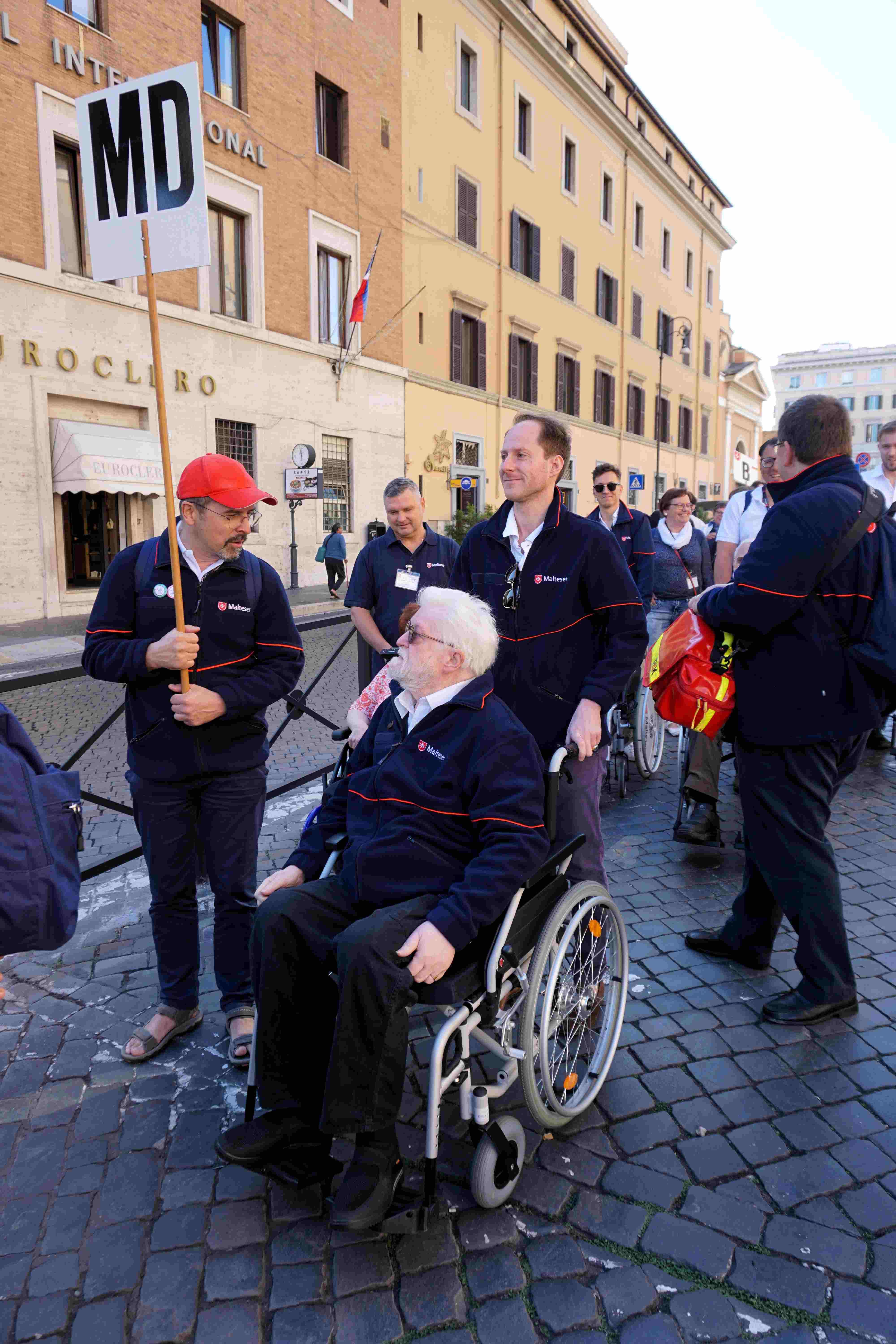 Person im Rollstuhl auf gepflastertem Platz, umgeben von mehreren Personen, eine hält ein Schild mit der Aufschrift 'MD'.
