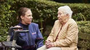 Seniorin in einer beigen Jacke sitzt neben einer Frau in Malteser Jacke auf einer Parkbank, vor den beiden steht ein Rollator