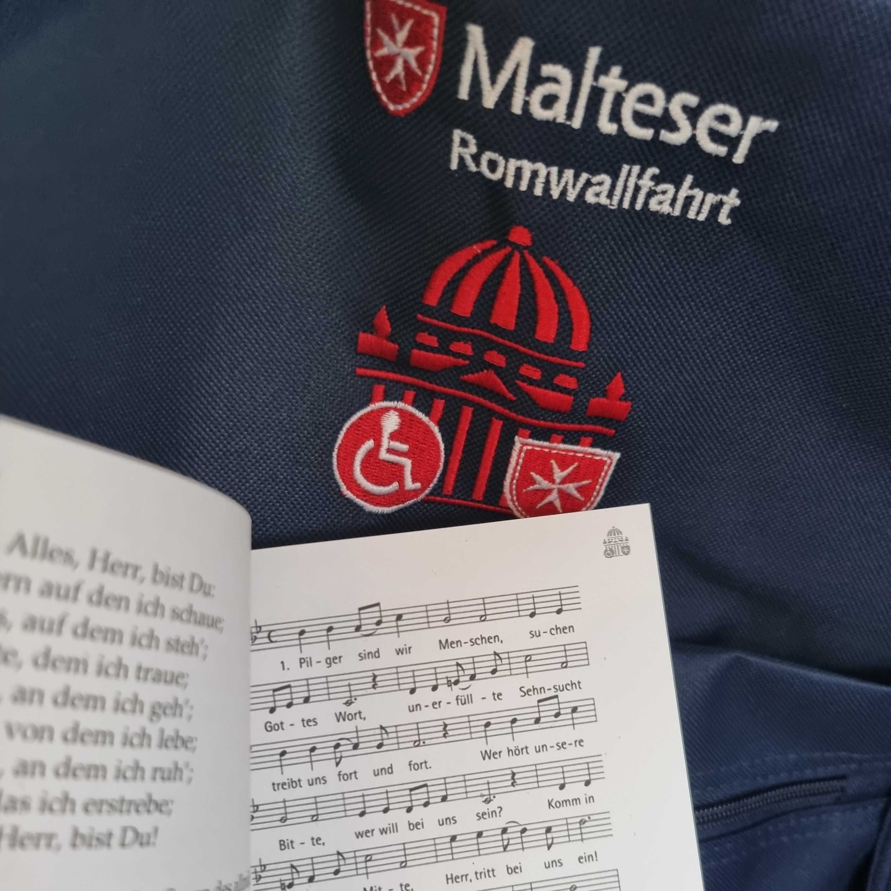 Rucksack mit rotem Malteser-Logo, Schriftzug 'Malteser Romwallfahrt' und Symbolen für Rollstuhl und Malteserkreuz, davor Notenblatt mit Liedtext.
