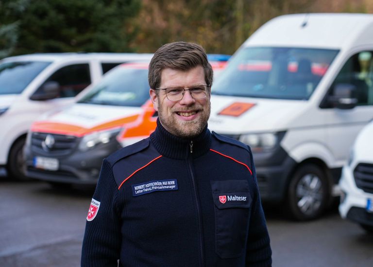 Ein Mann mit Bart in Malteser Jacke steht vor Fahrzeugen der Malteser.