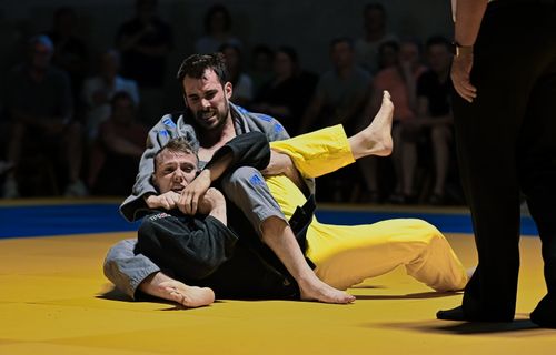 Zwei Judoka kämpfen am Boden, der obere Athlet im grauen Anzug setzt einen Würgegriff an, Kampfrichter am Mattenrand.