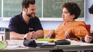 Ein Malteser Schulbegleiter sitzt mit einem Kind mit Behinderung am Tisch im Klassenszimmer,