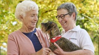 Eine Seniorin und eine ehrenamtliche Helferin der Malteser streicheln einen Hund