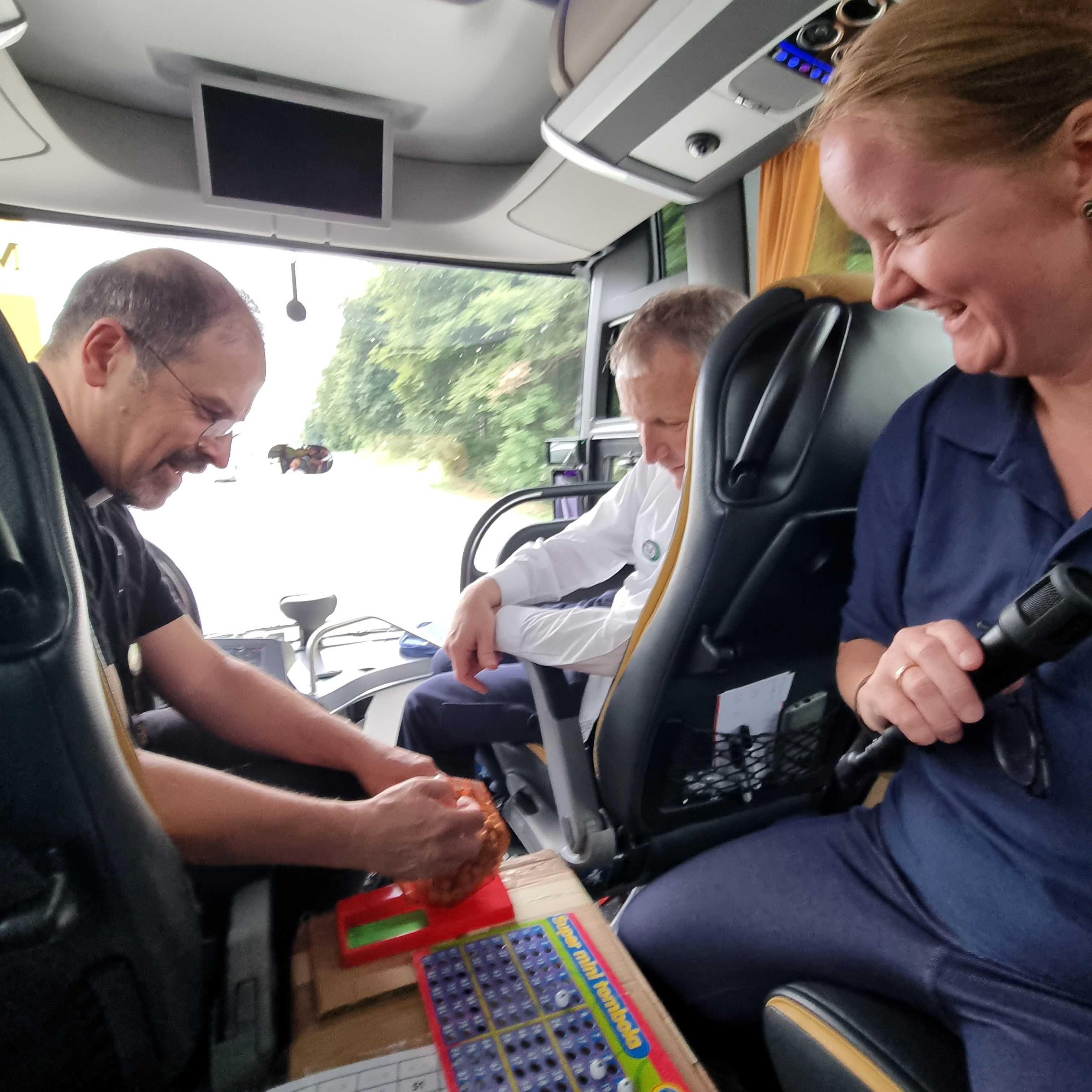 Drei Personen sitzen in einem Fahrzeug, eine hält ein Mikrofon, auf dem Tisch liegt ein Spiel mit bunten Spielfeldern und Kugeln.