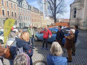 Eine Gruppe von Menschen steht im Freien auf einem gepflasterten Platz vor einem historischen Gebäude. In der Mitte spricht eine Person mit roter Jacke zu den Umstehenden. Hinter der Gruppe stehen mehrere geparkte Autos. Links im Bild sind ein gelbes Banner und bunte Luftballons zu sehen. Im Hintergrund reihen sich farbig gestaltete Altbauhäuser aneinander.