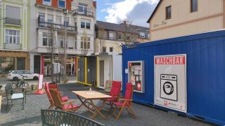 Ein blauer mobiler Container mit einem Banner „Waschbar“ steht auf einem Platz. Neben ihm stehen zwei weitere Container. Ein weißer und ein grauer. Vor den Containern sind Gartenmöbel aufgebaut.
