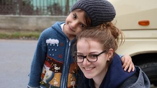 Junge Frau mit Brille wird von einem lächelnden Kind mit Mütze und bunter Strickjacke an der Schulter umfasst. Beide lächeln