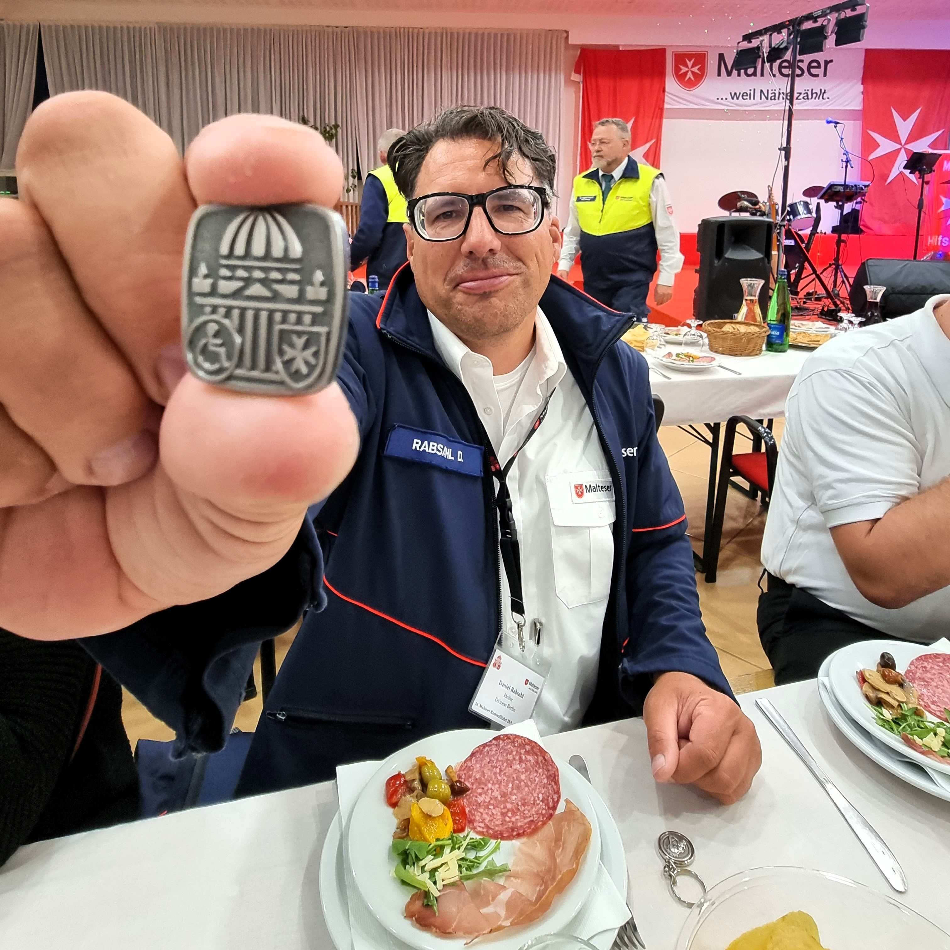Person hält eine Medaille der Malteser Romwallfahrt vor einem gedeckten Tisch mit Teller und Besteck in einem Saal mit weiteren Personen und roter Dekoration.
