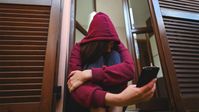 Eine junge Frau mit einer Kapuze über dem Kopf sietzt und schaut auf ein Smartphone 