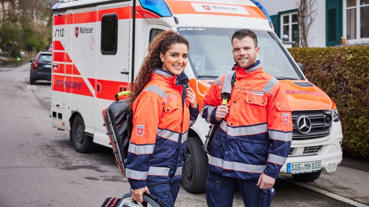 Rettungshelfer und Rettungshelferin der Malteser stehen vor einem Rettungswagen