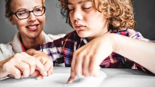 Frau sitzt hinter einem Jungen, der in Blindenschrift liest.