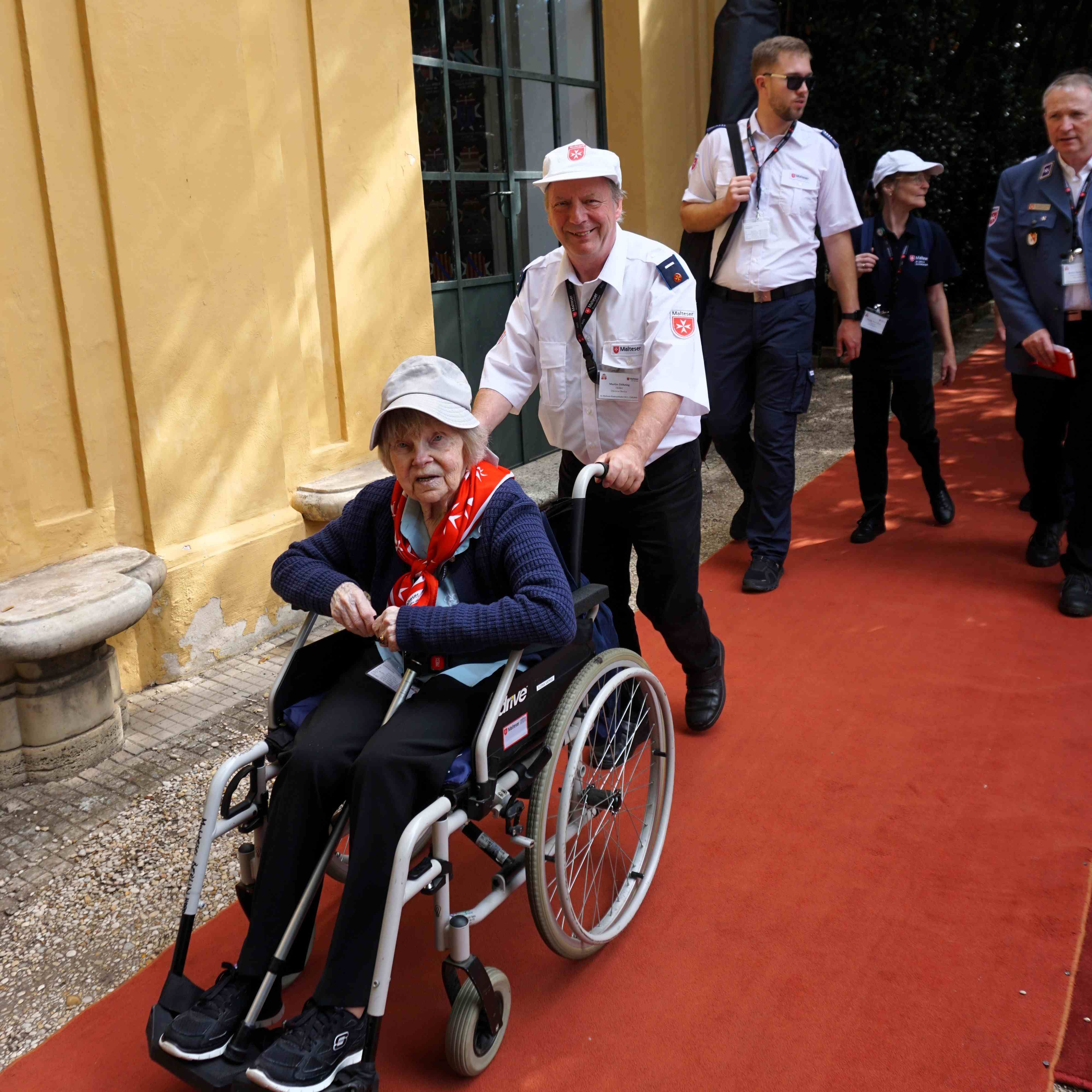 Person im Rollstuhl wird von einem Malteser Helfer auf rotem Teppich vor gelber Hauswand geschoben.