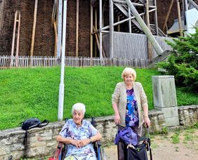 Eine Frau in einem Rollstuhl und eine Frau mit einer Gehhilfe stehen vor einem Gradierwerk.