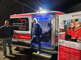Luis Moll und Dennis Lindemann präsentieren das Fahrzeug, rechts ist ein Roll-Up mit den Malteser-Standorten in Niedersachsen zu sehen, der Krankenwagen ist geöffnet und gibt den Blick auf den blau beleuchteten Innenraum frei