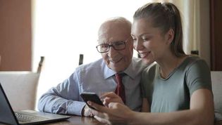 Junge Frau erklärt älterem Mann Smartphone und Laptop.