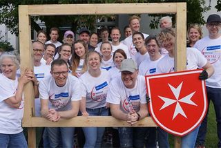 Menschen mit Firmentrikots blicken durch einen großen Bilderrahmen mit Malteserwappen