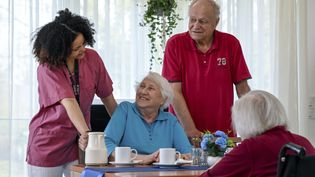 Pflegekraft in rotem Poloshirt steht an einem Tisch mit drei älteren Personen, zwei sitzen, eine im Rollstuhl, auf dem Tisch stehen Tassen, eine Kanne und eine Blumenvase.