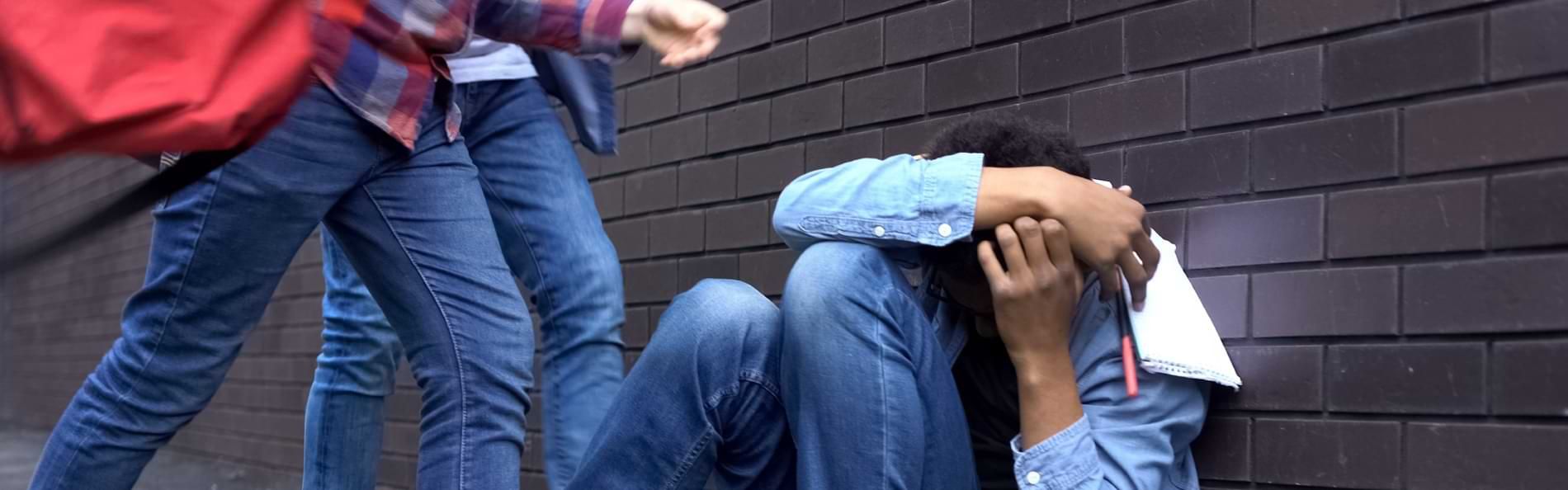 Ein Jugendlicher kauert an einer gemauerten Wand und hält schützend die Arme vor den Kopf; zwei weitere Personen stehen über ihm.