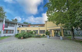 Ein modernes Gebäude mit Eingang, Hof und Bäumen unter blauem Himmel.