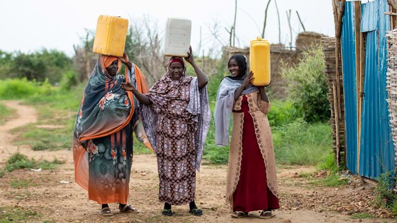 Drei Frauen tragen Wasserkanister auf einem Sandweg.