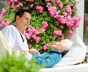 Ein junger Pfleger sitzt am Bett einer alten Frau. Das Bett steht auf der Terrasse.