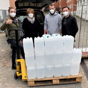 Vier Personen mit FFP2-Masken stehen hinter einer Palette mit Desinfektionsmittelkanistern vor Klinikgebäude und Fahrzeug.