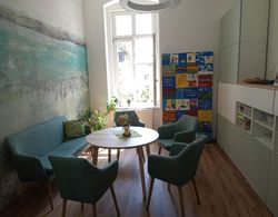 Runder Holztisch mit Blumen in Vase, umgeben von fünf grünen Sesseln und einer grünen Couch in hellem Raum mit Fenster und bunter Wandtafel.