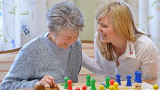 Zwei Frauen sitzen an einem Tisch und spielen ein Brettspiel mit bunten Spielfiguren und Würfeln.