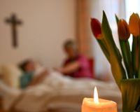Eine Kerze im Vordergrund. Im Hintergrund eine Ehrenamtliche am Bett im Gespräch mit einer Frau.am Bett einer 