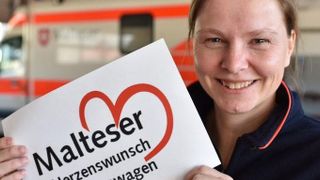 Malteser Mitarbeiterin zeigt das Logo des Herzenswunschkrankenwagens in die Kamera.