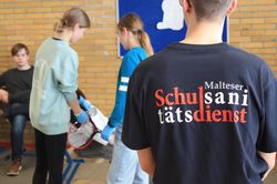 Junge von hinten mit großer Tshirt Aufschrift mit den Worten Malteser Schulsanitätsdienst. Vor ihm stehen zwei Schulsanitäterinnen, die einem fiktiven Patienten, der auf einem Stuhl sitzt, den Blutdruck messen.