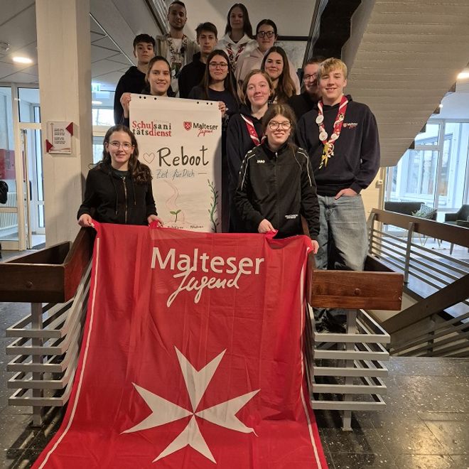 12 Jugendliche, die versetzt auf einer Treppe stehen und ein Banner der Malteser Jugend halten.