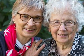 Malteser Mitarbeiterin legt Arm um ältere Frau und beide lachen in die Kamera.