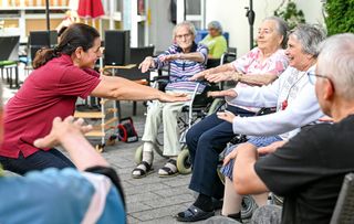 Ältere Menschen sitzen draußen in einer Reihe und strecken die Arme nach vorne als Übung