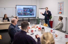 Der Malteser Jonas Gröger aus Gelsenkirchen steht vor einer Leinwand, auf der die Präsentation zum Projekt "Wohlfühlmorgen" zu sehen ist. Die übrigen Anwesenden hören vom Tisch aus zu.