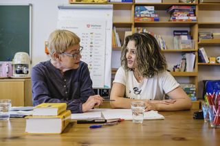 Zwei Frauen sitzen an einem Tisch vor einem Bücherregal und haben Bücher, Stifte und Zettel vor sich. Die linke Dame erklärt der rechten Dame etwas.