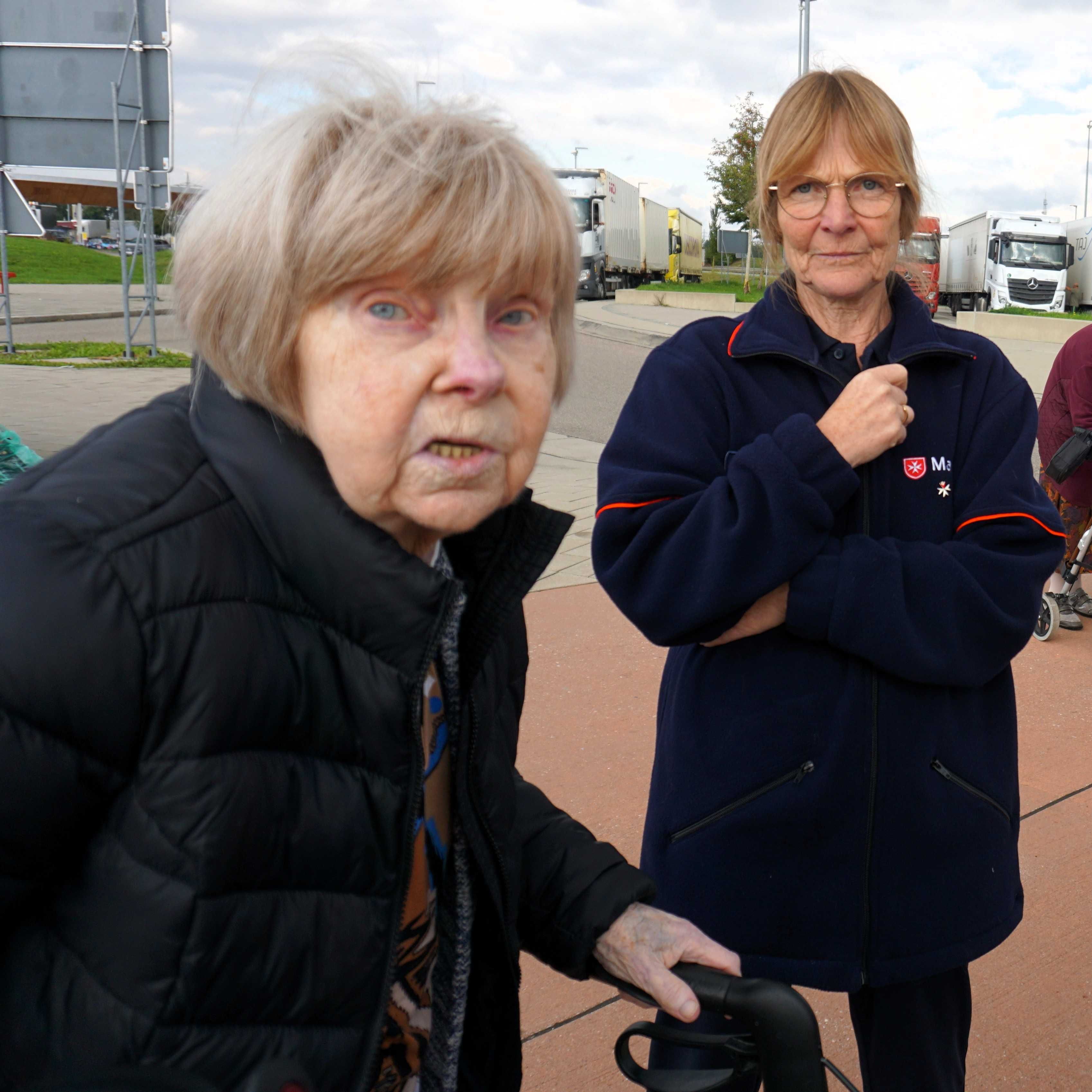 Zwei ältere Frauen, eine mit schwarzer Steppjacke und Rollator, die andere mit dunkelblauer Jacke mit rotem Streifen und Malteser Logo, stehen auf einem Parkplatz mit Lkw im Hintergrund.
